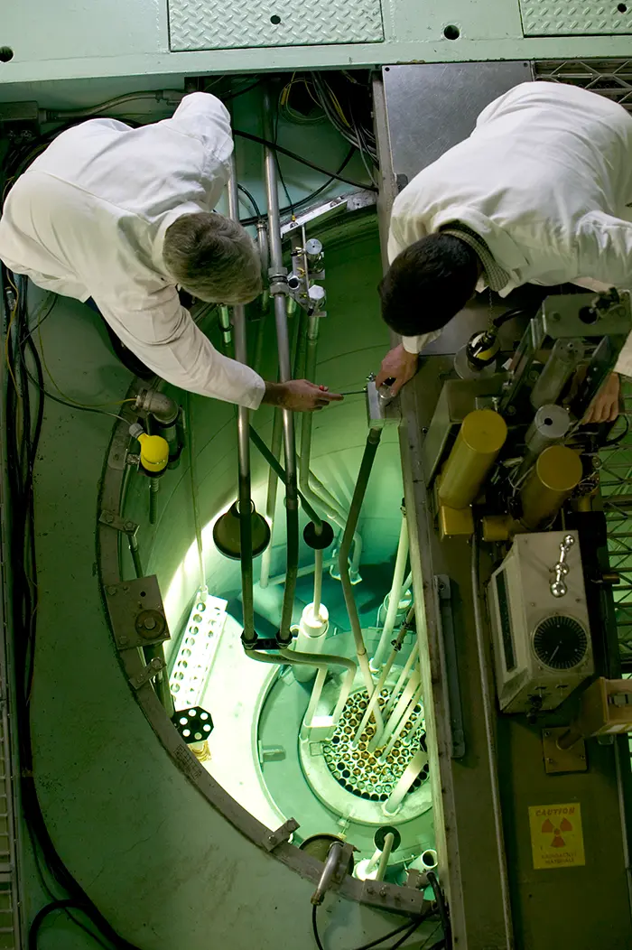 Two people in white lab coats lean over an open area with a glowing green light and instruments inside.